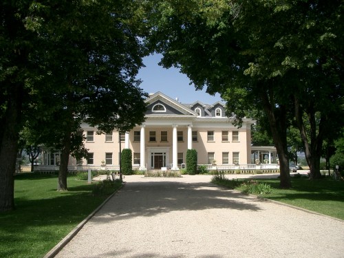 A large, elegant pink mansion with white columns, surrounded by green trees and a gravel pathway.