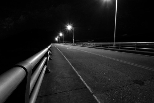A dimly lit road on a bridge at night, with streetlights illuminating the path and a railing along the side.