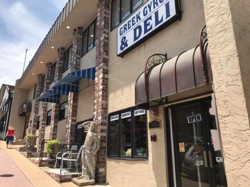 Exterior of a Greek Gyro & Deli with a statue outside, featuring an open sign and nearby shops.