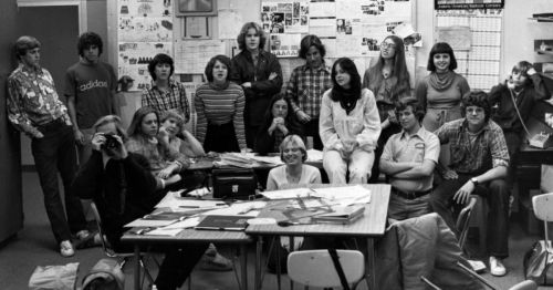 A black-and-white photo of a diverse group of students posing in a classroom, with desks and posters in the background.