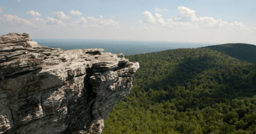 A rocky cliff overlooks a lush green valley under a blue sky with scattered clouds.