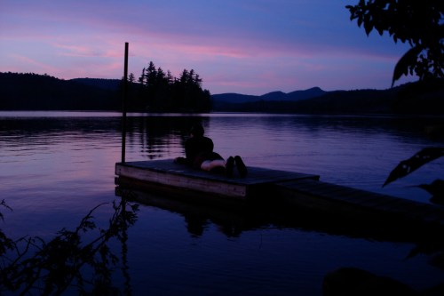 A serene lake at dusk with a person relaxing on a dock, surrounded by trees and mountains in the background.