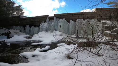 Frozen waterfall cascading over a stone dam, surrounded by snow and bare trees under a blue sky with clouds.