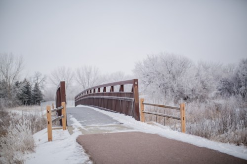 A snow-covered bridge spans a path through a winter landscape with frosted trees and a cloudy sky.