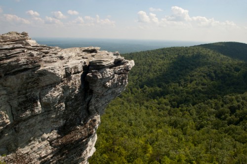 A rocky cliff edge overlooks a lush green valley and distant hills under a partly cloudy sky.