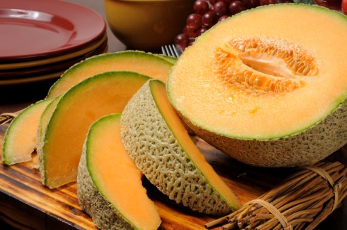 A halved cantaloupe with slices arranged on a wooden board, surrounded by grapes and plates in the background.