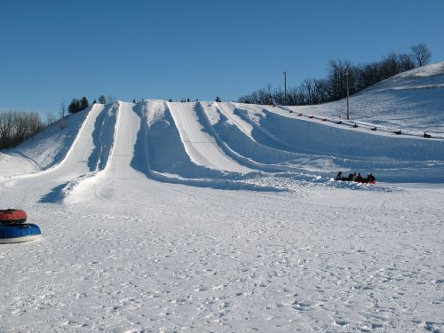 A snowy hill with multiple sledding lanes and people enjoying tubing on a clear winter day.