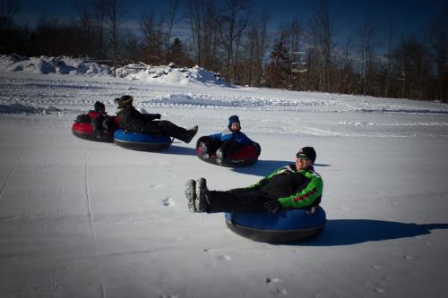 Four people sitting on snow tubes, sliding down a snowy hill on a clear winter day. Trees in the background.