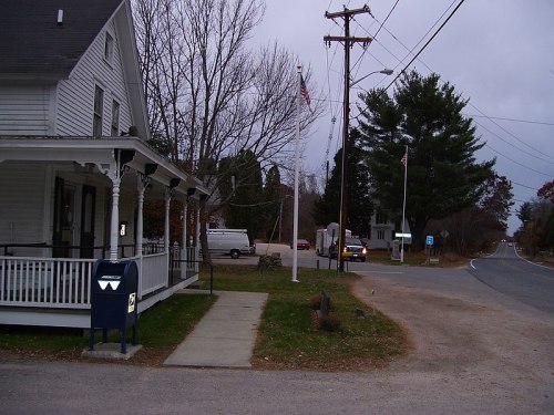 A quiet street scene featuring a white house, a mailbox, and a flagpole, with power lines and a road intersection nearby.