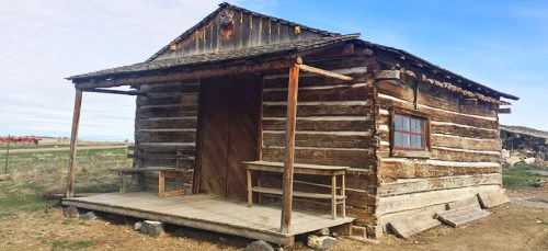 A rustic log cabin with a porch, set in an open landscape under a blue sky.