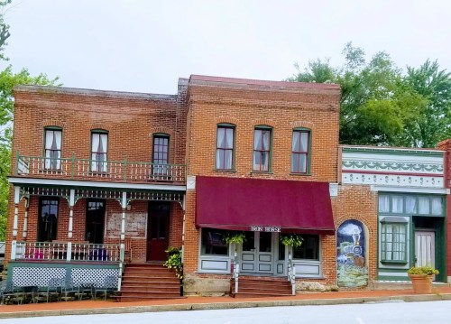 Historic brick buildings with colorful facades and a maroon awning, set against a backdrop of greenery.