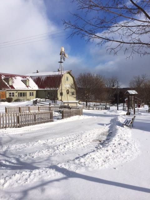 A snowy landscape featuring a yellow house, a windmill, and a wooden fence under a blue sky with clouds.