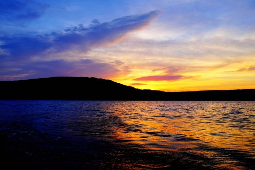 A vibrant sunset over a calm lake, with colorful reflections on the water and silhouetted hills in the background.
