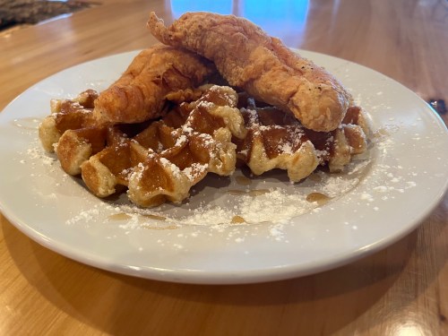 Plate of crispy fried chicken on top of golden waffles, dusted with powdered sugar and drizzled with syrup.