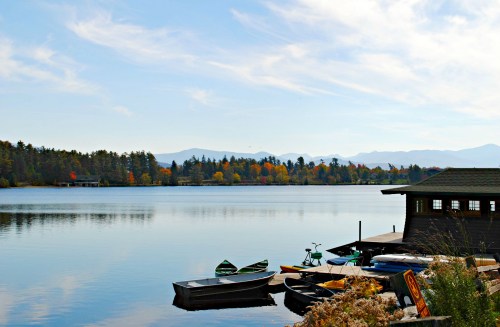 A serene lake scene with boats docked, surrounded by trees and mountains under a clear blue sky.