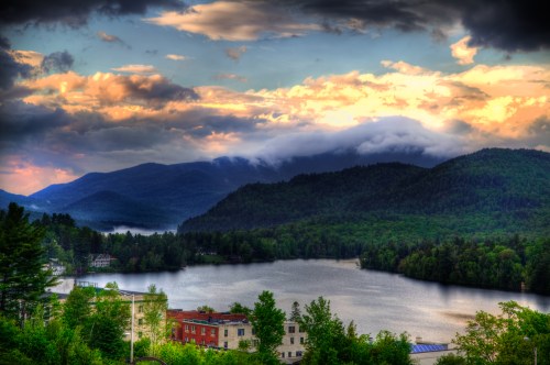 A scenic view of mountains and a lake under a cloudy sky, with lush greenery and a small town in the foreground.