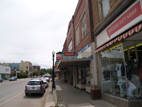 A street view of a small town featuring shops, parked cars, and a cloudy sky.