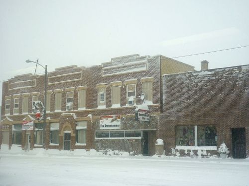 Snow-covered buildings line a street, with signs partially obscured by snow, creating a quiet winter scene.