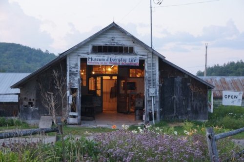 A rustic barn with a sign reading "Museum of Everyday Life," surrounded by flowers and greenery.