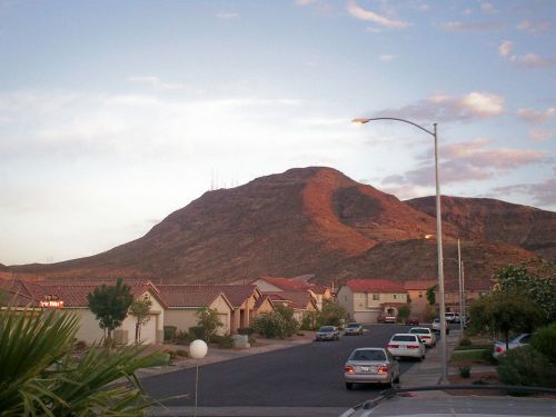 A residential neighborhood with houses and cars, set against a backdrop of a large, sunlit mountain.