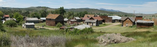 A panoramic view of a historic ghost town with wooden buildings surrounded by green fields and distant mountains.