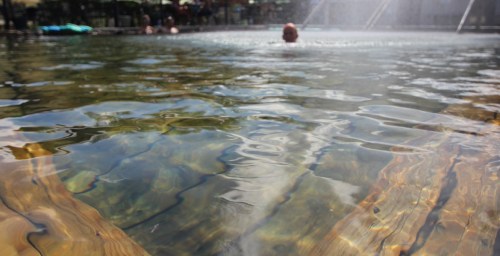 A serene pool scene with clear water reflecting wooden beams and people swimming in the background.