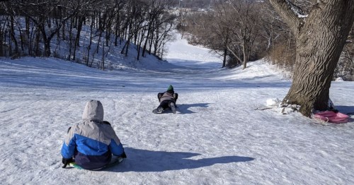 Two children sledding down a snowy hill, surrounded by trees, with a clear blue sky above.