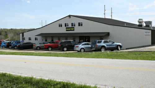 A large metal building with a sign, surrounded by parked cars, along a rural road on a sunny day.