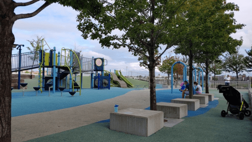 A colorful playground with slides and climbing structures, surrounded by trees and benches, with a stroller nearby.