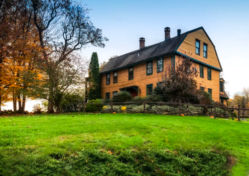 A charming two-story wooden house surrounded by trees and a green lawn, with autumn foliage in the background.