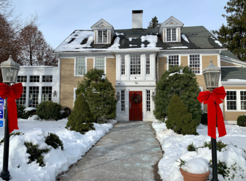 A large, snow-covered house with a red door, festive wreath, and holiday decorations on the front yard.