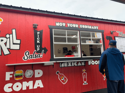 A person stands in front of a vibrant red food truck with a menu and signs for Mexican food and salsa.