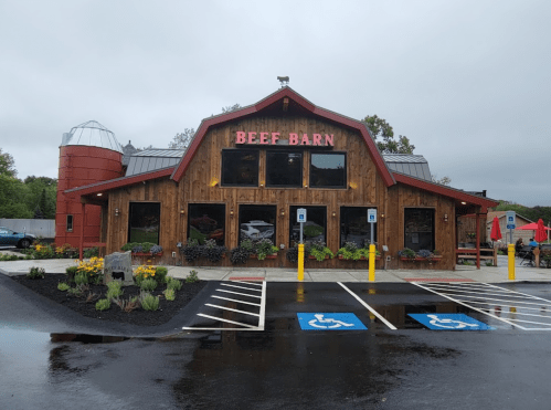 A rustic wooden building with "BEEF BARN" signage, surrounded by flowers and a silo, on a rainy day.