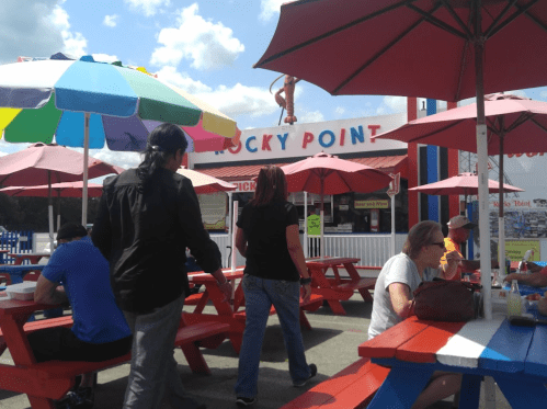 People walking past colorful umbrellas and picnic tables at a casual outdoor dining area called "Rocky Point."