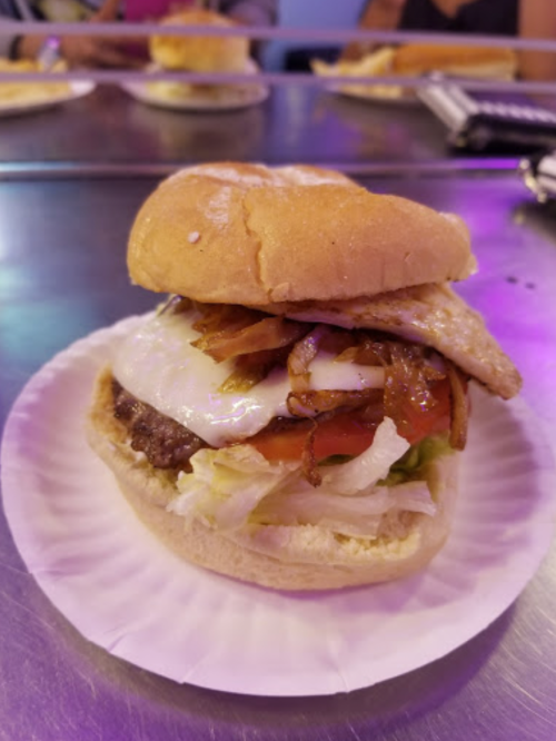 A close-up of a burger with a beef patty, fried egg, lettuce, tomato, and grilled onions on a paper plate.