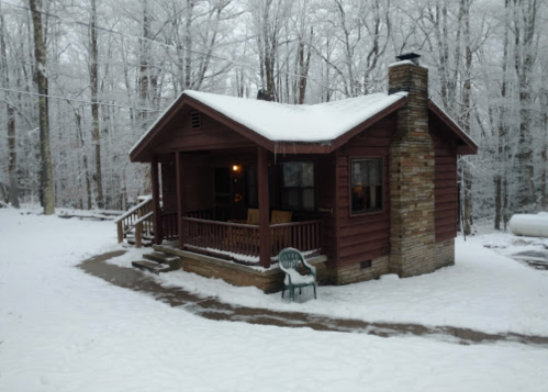 A cozy cabin surrounded by snow-covered trees, with a porch and stone chimney, in a winter landscape.