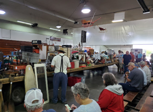 A busy indoor auction with people seated and browsing various items on display at a long table.