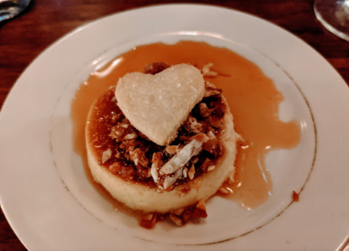 A creamy dessert topped with crushed nuts and a heart-shaped cookie, served on a plate with caramel sauce.