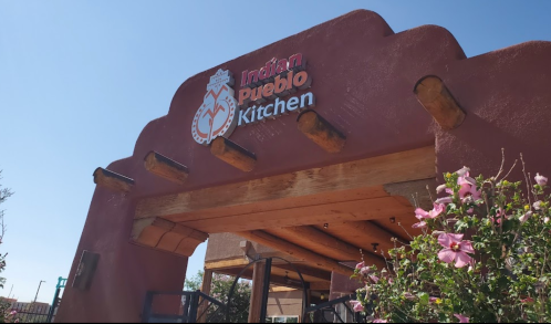 Sign for Indian Pueblo Kitchen, featuring a rustic adobe-style entrance with wooden beams and pink flowers.