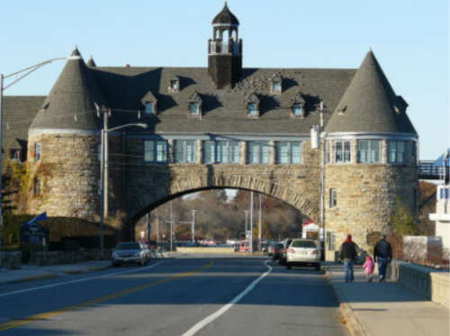 Historic stone building with a tower, featuring an archway over a road, surrounded by trees and parked cars.