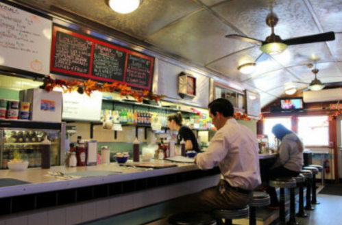 A cozy diner interior with patrons at the counter, colorful menus, and autumn decorations.