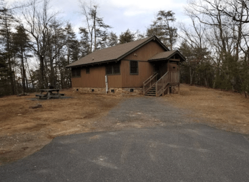 A brown wooden cabin surrounded by trees, with a gravel path and picnic table in front.