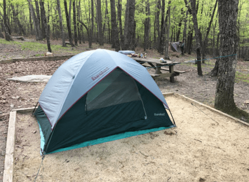 A gray and green tent set up in a wooded campsite with a picnic table and scattered camping gear in the background.