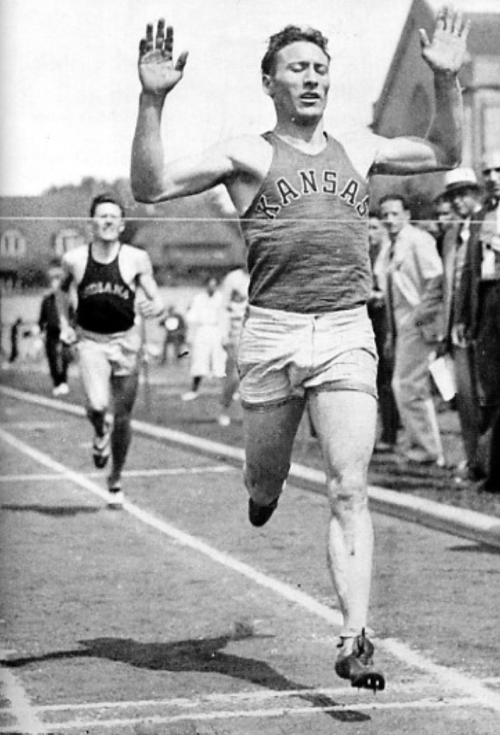 A runner in a Kansas tank top crosses the finish line, celebrating victory, with another competitor behind him.