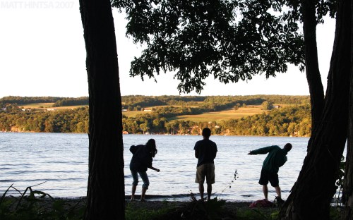 Silhouettes of four people by a lake at sunset, surrounded by trees and a scenic landscape in the background.