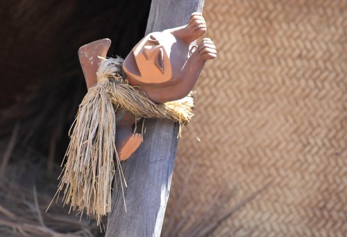 A small clay figure with a triangular face and straw adornment, attached to a wooden post against a woven background.