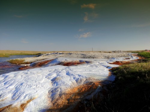 A colorful geothermal landscape with white mineral deposits and a flowing stream under a clear sky.