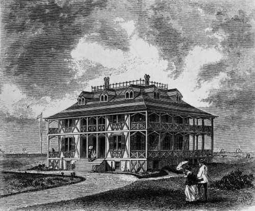 An ornate two-story building with a porch, surrounded by greenery, featuring people sitting outside.