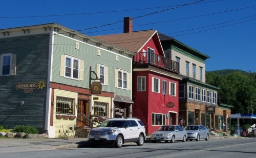 Colorful buildings line a street, featuring shops and a café under a clear blue sky. Cars are parked along the road.