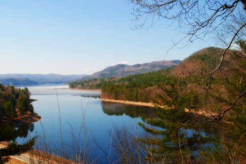 A serene lake surrounded by mountains and trees under a clear blue sky. Reflections shimmer on the water's surface.